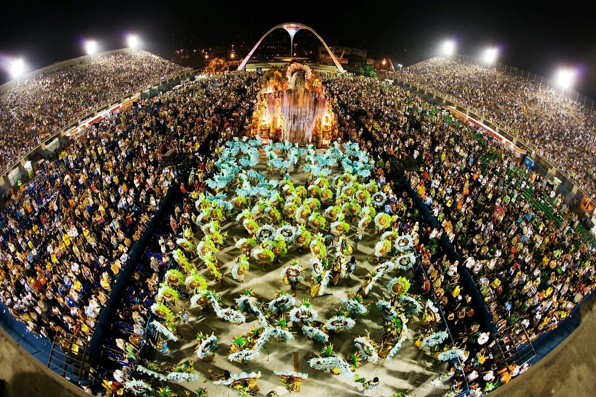Vista aérea noturna do Sambódromo com multidões e passistas em fantasias elaboradas de plumas, sob holofotes dourados e azuis.